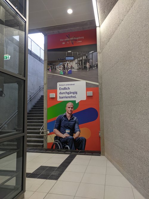 Ein Plakat im Bahnhof auf welchem ein Mensch mit Rollstuhl ist. Mit dem Text: Einfach verbindend: Endlich durchgängig barrierefrei.